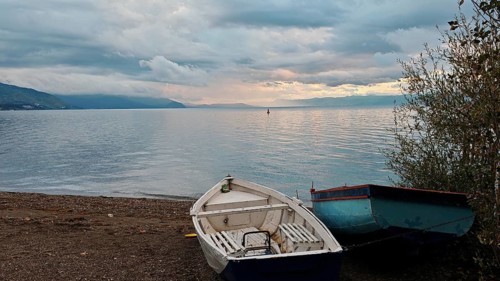 Two abandoned boats on the shore of a lake with mountains in the background under a cloudy sky,