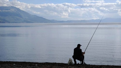 An angler sits on the shore of a peaceful lake, surrounded by mountains and a peaceful, natural