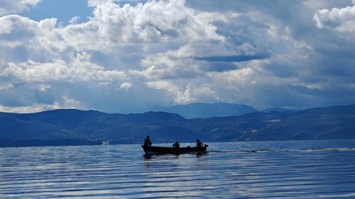 A small boat with silhouettes in a calm lake surrounded by mountains and dramatic cloud formations,