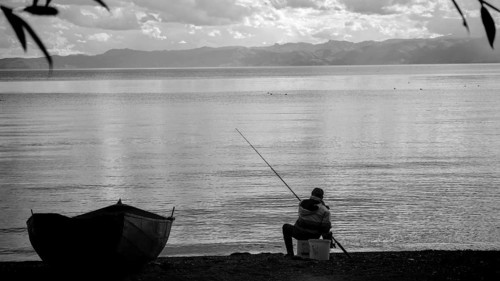 An fisherman sitting on the shore of a calm lake, with a small boat and a black and white