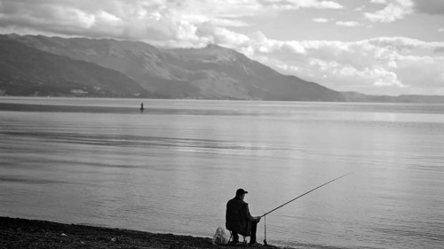 Angler sitting on quiet lakeside with views of mountains on horizon, Ohrid Lake, North Macedonia