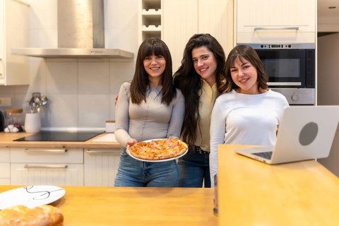 Three young women smiling and looking at camera while one is holding a freshly baked pizza in a