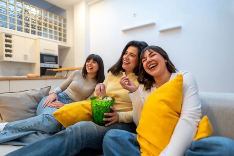 Three young female friends sitting on a sofa in a modern apartment, enjoying an entertaining movie