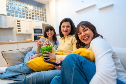 Three young women laughing and relaxed on a couch at home, enjoying a cozy movie night together