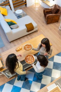 Three happy young women gathered around a wooden kitchen island sharing homemade pizza, chatting