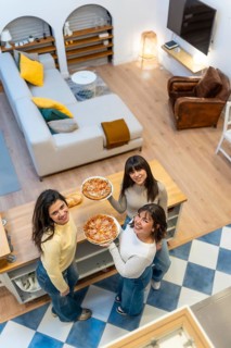 Three cheerful young women standing in an open plan apartment, holding pizzas and looking up while