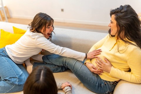Two cheerful women expressing joy and support, one touching the pregnant belly of the other,