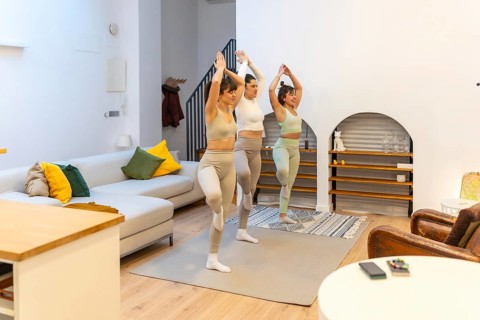 Three young women practice tree pose together on yoga mats in a modern apartment living room,