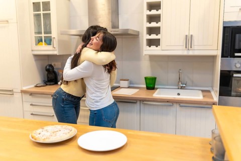 Two smiling adult women sharing a warm embrace in a bright, contemporary kitchen, expressing joy