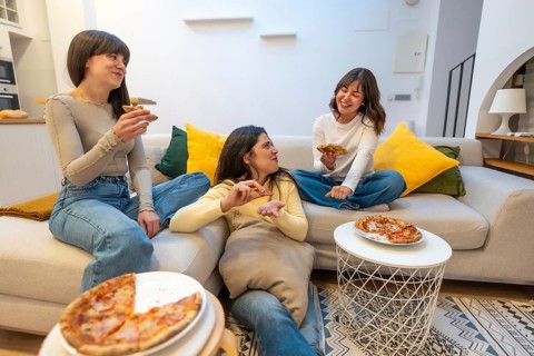 Three young women laughing and talking while eating pizza slices on a sofa in a modern living room,