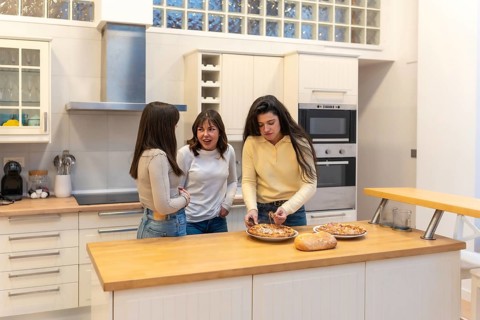 Three young women in a modern kitchen preparing homemade pizza, chatting and laughing while bonding