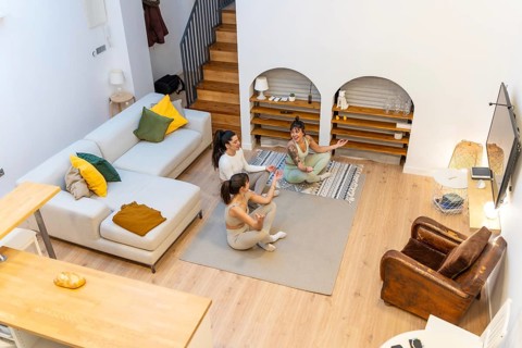 Three young women sitting in a living room, smiling and relaxing on yoga mats, enjoying a peaceful