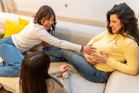 Two friends sharing a tender moment, sitting on a sofa and happily touching the belly of a smiling
