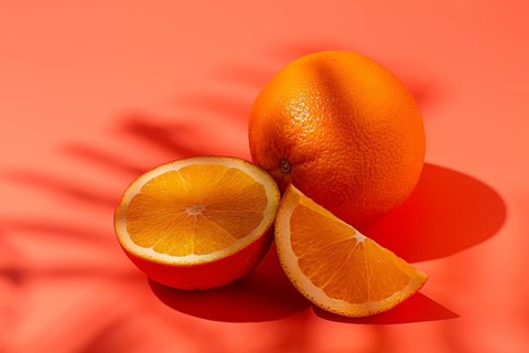 On the coral surface, next to two neatly sliced slices, there is a whole orange. Sunlight casts