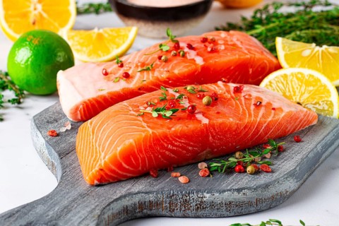 Two pieces of salmon fillet sit on a wooden cutting board. They are topped with herbs and pink
