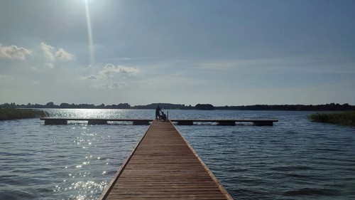 A long wooden walkway leads across a calm lake under bright sunlight, Masuria, Poland