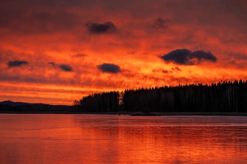 A dramatic sunset over a still, frozen lake and forest with orange-red hues in a cloudy sky,
