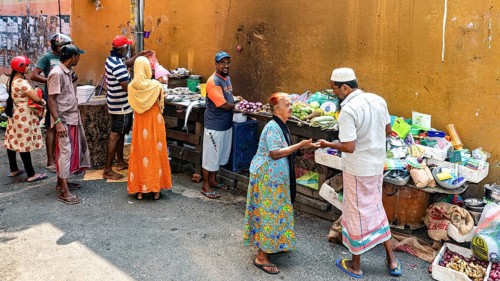 People trade and interact at a market stand full of fresh vegetables and goods