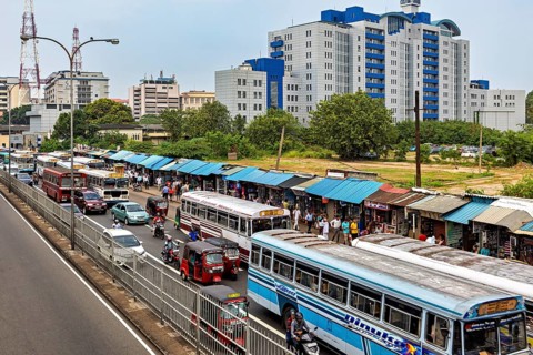 City street with several buses and market stalls, surrounded by large buildings and trees, buses in