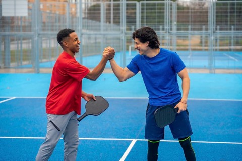 Two diverse men playing pickleball are celebrating with a high five on a blue outdoor court,