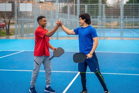 Two smiling male pickleball players of different ethnicities shaking hands on a vibrant blue court