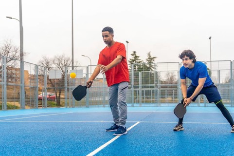 Two young men play pickleball on a bright blue outdoor court, one about to hit a yellow ball with