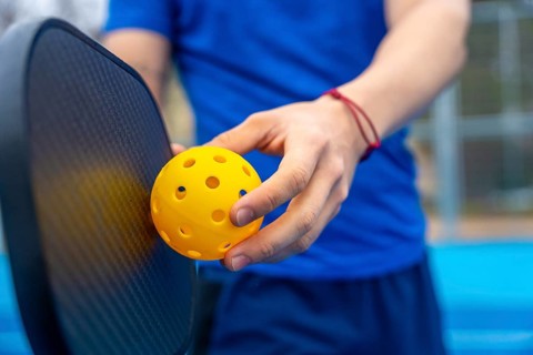Hands holding a yellow pickleball and paddle on an outdoor court, poised to serve or dinkclose up