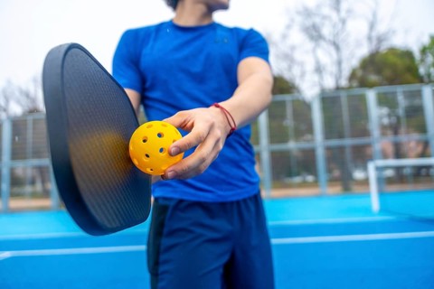 Athlete preparing to serve, holding a yellow pickleball and paddle on a vibrant blue outdoor court,