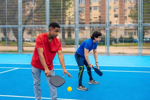Young men poised on a bright blue court with paddles and ball, ready for an outdoor pickleball