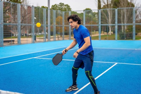 Young man playing pickleball on a blue outdoor court, striking a yellow ball with a black paddle,