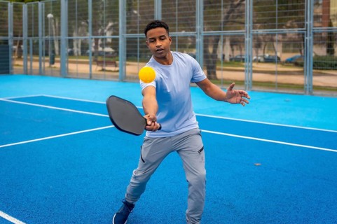 Young man strikes a yellow pickleball with a paddle on a vibrant blue outdoor court, showcasing