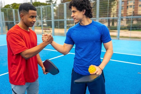 Two young diverse male athletes shaking hands on a blue pickleball court, holding paddles and a