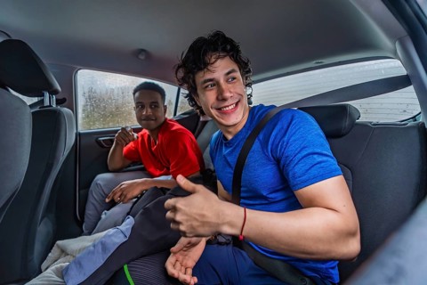 Two young diverse men sitting in the back seat of a car, one smiling and giving a thumbs up,