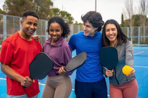 Group of diverse young friends smiling and embracing, holding pickleball paddles and a ball on a