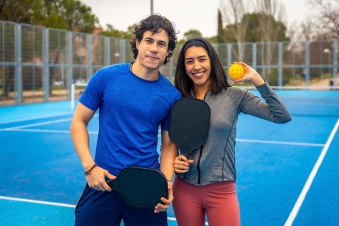 Enthusiastic young couple standing on a blue pickleball court, smiling and holding paddles and a