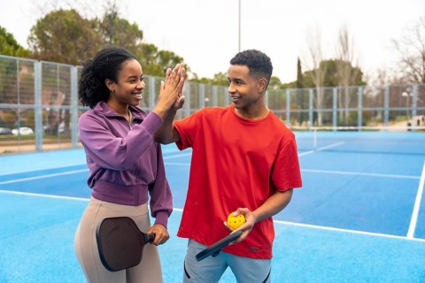 Young diverse couple playing pickleball on a blue court, celebrating with a high five, showing