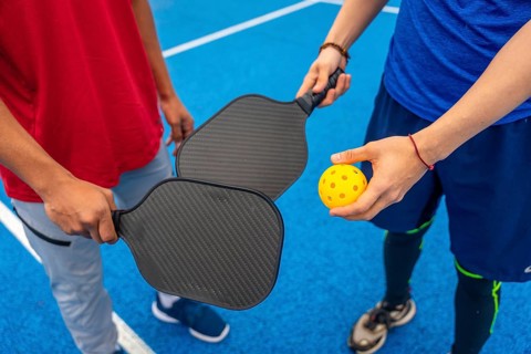 Two people are ready to play pickleball, holding paddles and a bright yellow wiffle ball on a