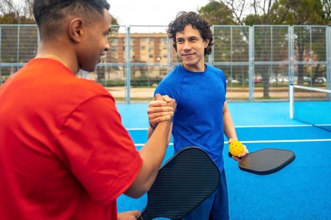 Two diverse male pickleball players shaking hands on a blue court, proudly displaying their paddles