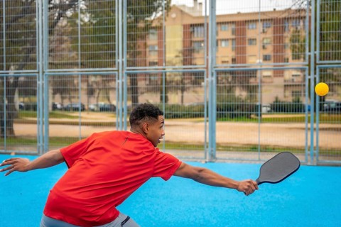 Young man in a red top lunging with paddle to hit a yellow pickleball on a bright blue outdoor