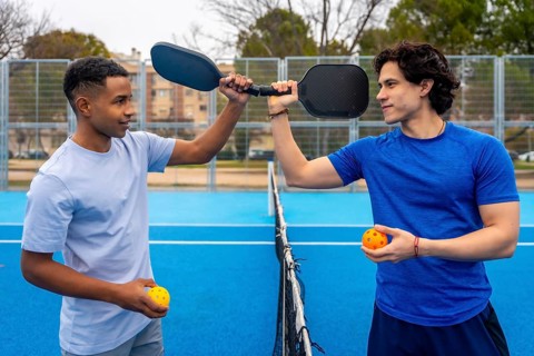 Two men stand on an outdoor pickleball court holding paddles and balls, sharing a friendly moment