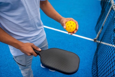 Person standing on a bright blue pickleball court, holding a black paddle and a yellow perforated