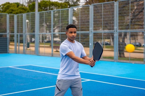 Young man poised to strike a yellow ball with a paddle on an outdoor blue pickleball court, focused