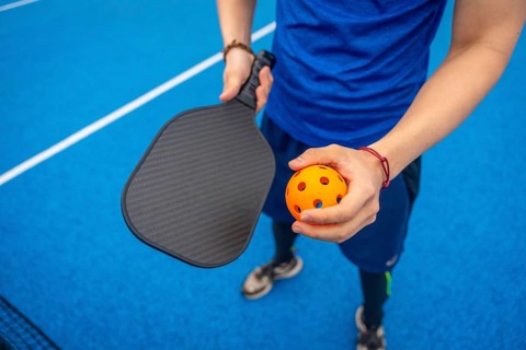 Player standing on a vibrant blue pickleball court, holding a textured paddle and an orange wiffle