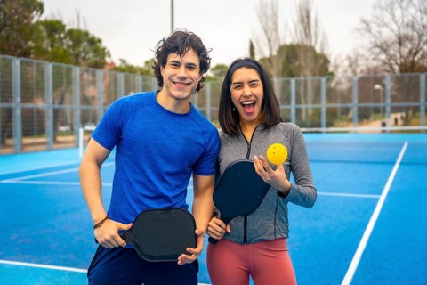Two young athletes smiling on a bright blue outdoor pickleball court, holding paddles and a ball,