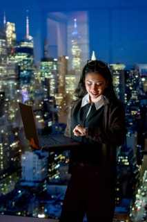 Young businesswoman standing by a large window, working on a laptop and checking her smartwatch