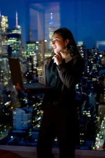 Young businesswoman standing confidently by a large window with her laptop, looking out at the
