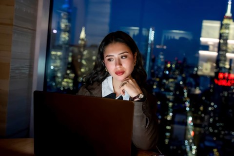Young businesswoman working late on a laptop in a modern office, gazing toward the illuminated