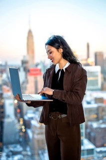 Young businesswoman standing by a window, focused on her laptop while working in an urban setting