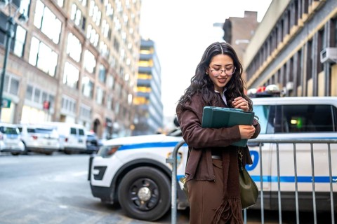 Young executive woman is holding a modern tablet and smiling, standing outdoors in a bustling city