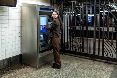 Young businesswoman with glasses and dark wavy hair smiling while interacting with a ticket machine